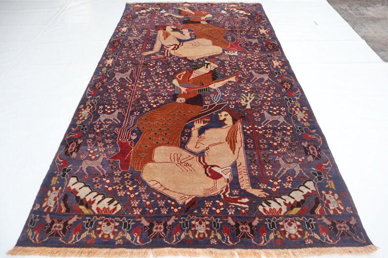 May include: A rectangular, hand-woven rug with a deep blue background and intricate patterns. The rug features figures in beige tones, deer, and floral motifs. The border is a mix of red, beige, and blue, with a fringed edge.