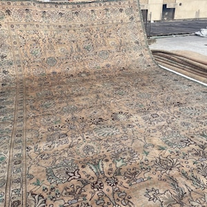 May include: A large, rectangular, antique rug with an intricate floral pattern in beige, brown, and teal. The rug has a detailed border and is partially rolled up, showcasing its size and design. The rug is likely made of wool.