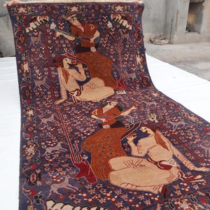 May include: A detailed Persian rug with a deep blue background and intricate patterns. The rug features figures in traditional attire, deer, and floral motifs in shades of red, beige, and brown. The rug is displayed on a white surface.