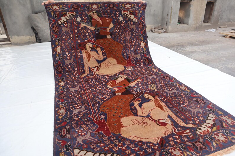 May include: A detailed Persian rug with a deep blue background and intricate patterns. The rug features figures in traditional attire, deer, and floral motifs in shades of red, beige, and brown. The rug is displayed on a white surface.