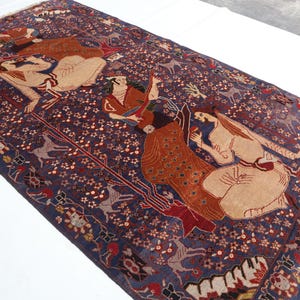 May include: A rectangular, hand-woven rug with a dark blue background and intricate patterns. The rug features figures in beige, red, and brown, along with floral and animal motifs. The rug has a fringed edge.