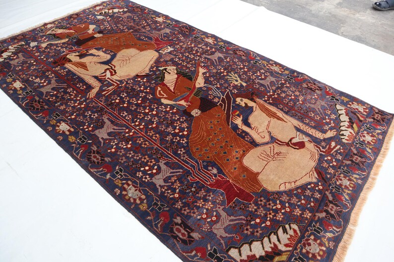 May include: A rectangular, hand-woven rug with a dark blue background and intricate patterns. The rug features figures in beige, red, and brown, along with floral and animal motifs. The rug has a fringed edge.
