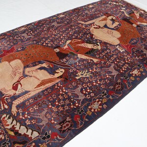 May include: A rectangular, antique Persian rug with a deep blue background and intricate patterns. The rug features figures in tan, red, and brown, with floral and geometric designs. The edges have a fringed border.