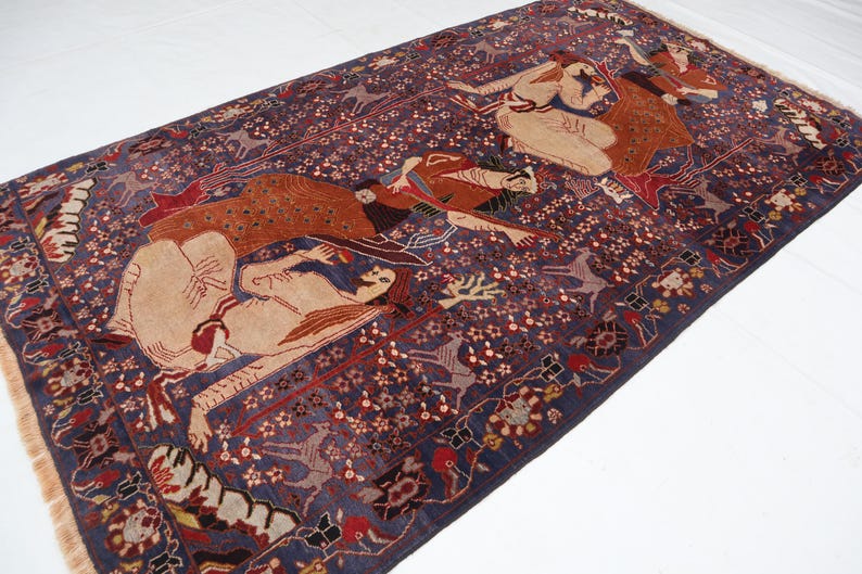 May include: A rectangular, antique Persian rug with a deep blue background and intricate patterns. The rug features figures in tan, red, and brown, with floral and geometric designs. The edges have a fringed border.
