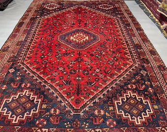 May include: A rectangular area rug featuring a deep red central field with a large navy blue diamond-shaped medallion. The rug is decorated with intricate geometric patterns, floral motifs, and stylised bird figures in cream, blue, and red.