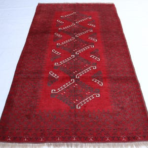 Pode incluir: Um tapete retangular, tecido à mão, com um campo vermelho escuro e um design central de padrões geométricos repetidos em verde escuro e branco. O tapete tem franjas em ambas as extremidades e é adequado para uma sala de estar ou quarto.