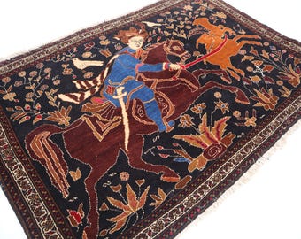 Puede incluir: Una alfombra de diseño intrincado con un guerrero a caballo, en tonos azules, marrones y naranjas sobre un fondo oscuro. La alfombra muestra motivos florales y foliares, con un borde detallado. Las dimensiones de la alfombra no son visibles.