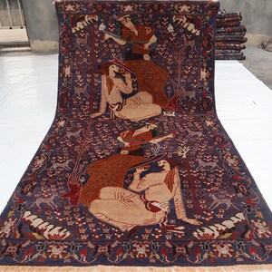 May include: A large, rectangular rug with a dark blue background and intricate patterns. The rug features figures, animals, and floral motifs in shades of brown, red, and beige. The rug has a fringed edge.