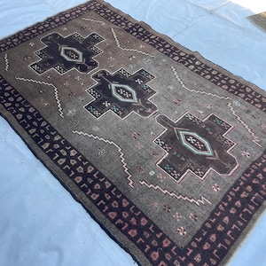 May include: A rectangular, hand-woven rug with a central design of three dark brown geometric medallions on a gray background. The rug has a dark brown border with repeating patterns and white zigzag accents.