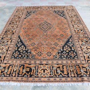 May include: A rectangular area rug featuring a central orange field with a dark navy blue diamond medallion and intricate geometric patterns. The border displays a repeating floral motif in orange, navy blue, and light gray with white fringe.