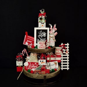 May include: A tiered wooden tray decorated for Christmas with red and white snowman figurines, candy canes, a red toy truck, and signs that say "Let it Snow", "Milk for Santa", "Believe", and "Merry & Bright".
