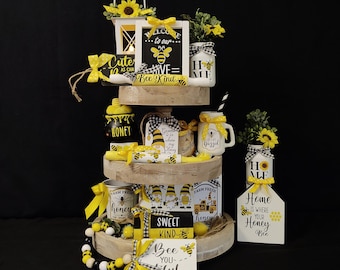 May include: A three-tiered wooden tiered tray decorated with a bee theme. The tray features a variety of bee-themed decorations, including signs, jars, and figurines. The signs include phrases like "Welcome to our Hive", "Sweet Kind", "Bee You-tiful", and "Home is where your Honey Bee".