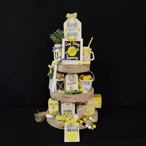 May include: A three-tiered wooden tray decorated with a lemon theme. The top tier features a sign that says "Home Sweet Home", a chalkboard sign that says "Easy Peasy Lemon Squeezy", and a mason jar with a straw. The middle tier features a lantern, a sign that says "Lemons Lemonade", a small bucket with lemons, and a sign that says "Lemons 5c". The bottom tier features a sign that says "Squeeze Me", a box of lemons, a sign that says "Squeeze the Day", and a string of black and white beads with yellow lemon charms.