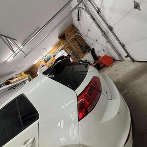 Puede incluir: Un coche blanco con portón trasero estacionado dentro de un garaje. El coche tiene ventanas tintadas de negro y luces traseras rojas. La puerta del garaje blanca está parcialmente abierta, revelando el interior con herramientas y equipos.