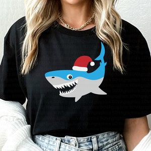 以下が含まれることがあります： サンタの帽子をかぶったサメのグラフィックがプリントされた黒いTシャツ。サメは青とグレーで、白い歯をしています。