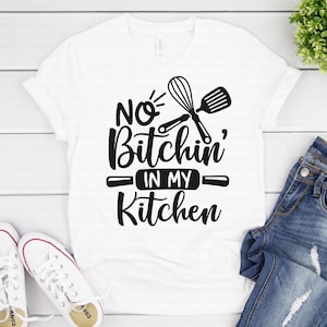 Peut inclure: T-shirt blanc avec l'inscription "NO Bitchin' IN MY Kitchen" en noir, en gras, dans une police manuscrite. Le motif comprend un fouet, une spatule et un rouleau à pâtisserie. Une paire de baskets blanches et un jean bleu sont à côté du t-shirt.