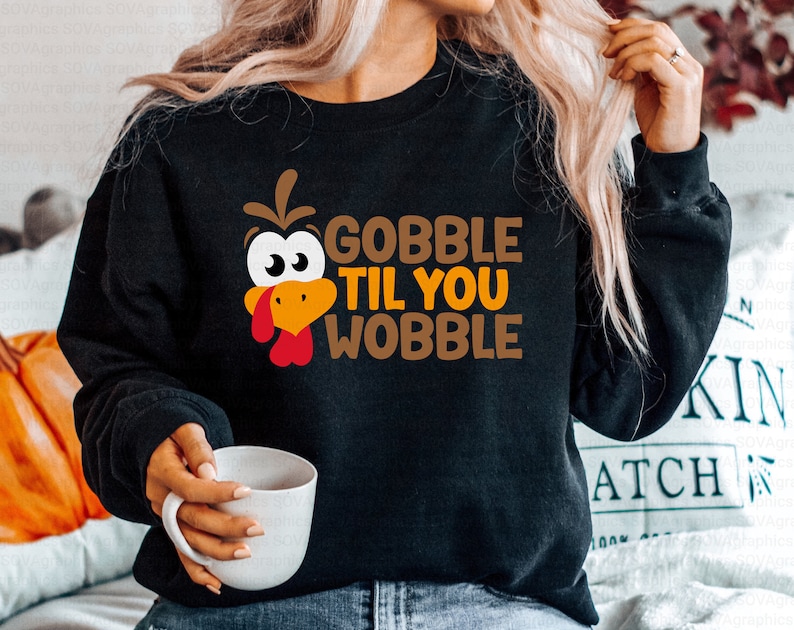 Może przedstawiać: Czarny sweter z kresk&oacute;wkową twarzą indyka i tekstem "Gobble Til You Wobble" w pomarańczowych i brązowych literach.