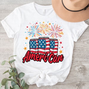 May include: White t-shirt featuring a patriotic design. The graphic includes three beverage cans decorated with the American flag, fireworks, and the word "American" in red script. The design is perfect for celebrating Independence Day.