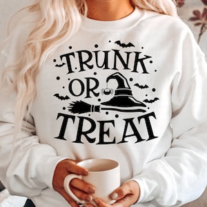 Op de afbeelding: Wit sweatshirt met zwarte tekst die "TRUNK OR TREAT" luidt met een grafische afbeelding van een heksenhoed en een bezem.