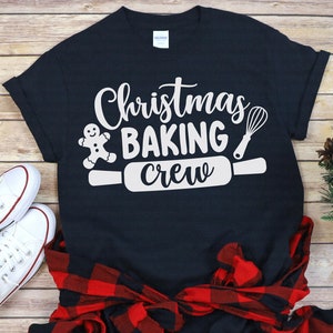 Può includere: Una maglietta nera con un design bianco che dice "Christmas Baking Crew". Il design include un omino di pan di zenzero, un mattarello e una frusta.