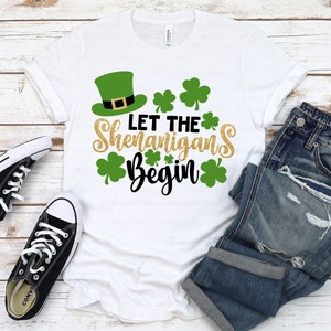 May include: White t-shirt with a green leprechaun hat and gold glitter text that reads "Let the Shenanigans Begin".