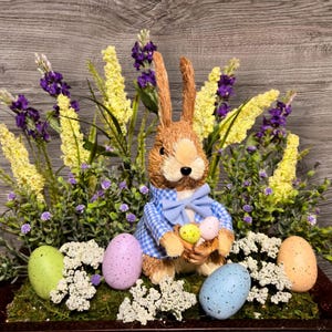 Könnte beinhalten: Ein braunes Kaninchen, das ein blau-weiß kariertes Hemd und eine blaue Schleife trägt, hält einen Korb mit bunten Ostereiern. Das Kaninchen ist von gelben und violetten Blumen sowie weißen Blumen umgeben.
