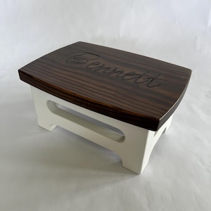 May include: A small wooden step stool with a dark brown top and white base. The top is engraved with the name "Bennett" in a cursive font. The stool has a rectangular top and a cut-out handle on each side.