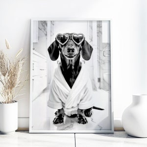 May include: A black and white photo of a dachshund wearing heart-shaped sunglasses and a white bathrobe. The dog is sitting on a white tiled floor.