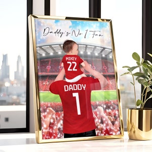 May include: A red and white soccer jersey with the word "Daddy" and the number "1" on the back. A smaller red and white soccer jersey with the word "Mikey" and the number "22" on the back is being held by the person wearing the larger jersey. The background is a blurred image of a soccer stadium with a crowd of people.