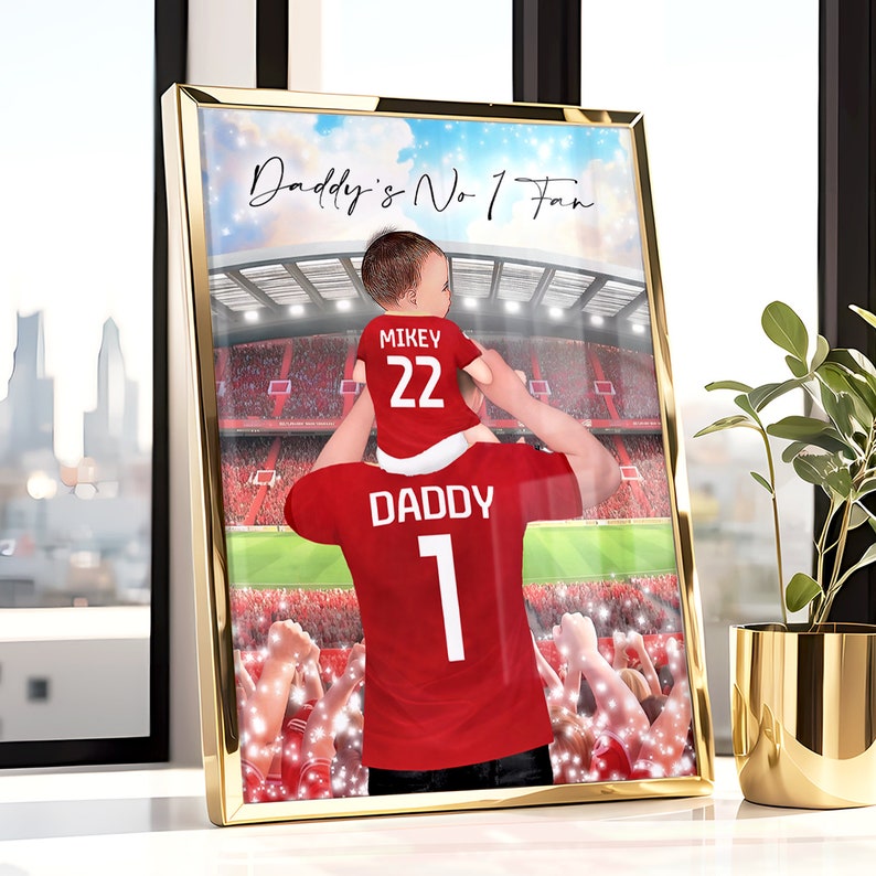 May include: A red and white soccer jersey with the word "Daddy" and the number "1" on the back. A smaller red and white soccer jersey with the word "Mikey" and the number "22" on the back is being held by the person wearing the larger jersey. The background is a blurred image of a soccer stadium with a crowd of people.