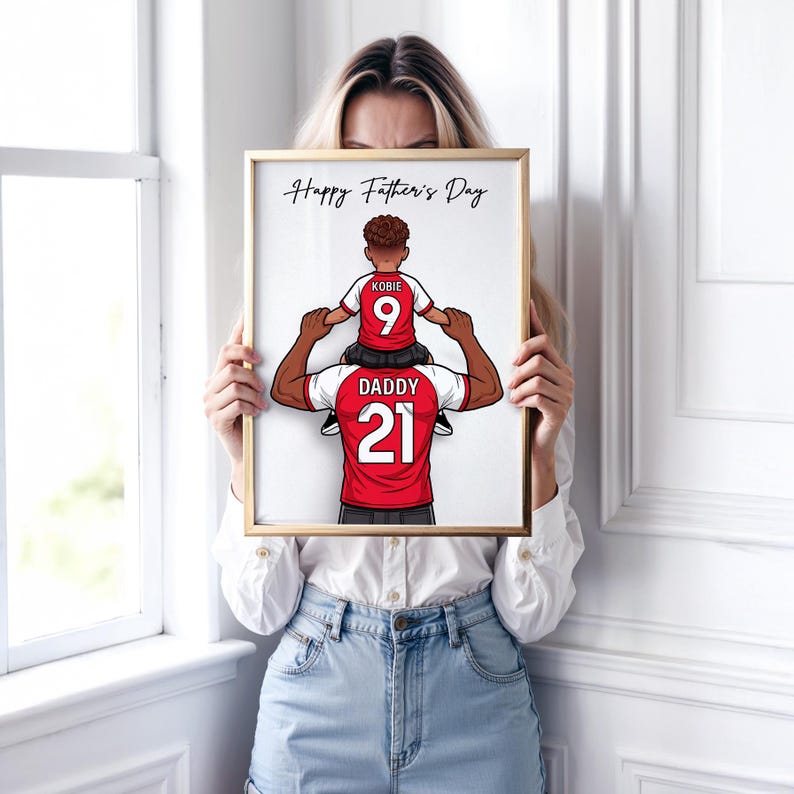 Pu&ograve; includere: Stampa incorniciata con bordo dorato. L'illustrazione mostra un padre e suo figlio, entrambi con maglie da calcio rosse e bianche. Il bambino &egrave; seduto sulle spalle del padre. Il testo in alto dice "Happy Father's Day."