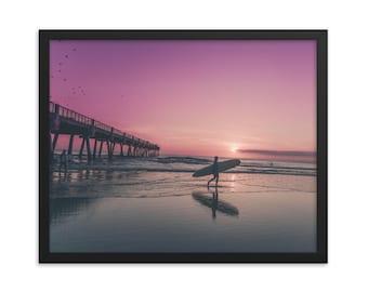 May include: A silhouette of a person walking on a beach with a surfboard under their arm. The beach is at sunset with a pink and purple sky. A pier is in the background.