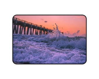 May include: A close-up of a wave crashing on the shore with a pier in the background. The sky is a vibrant pink and orange, creating a beautiful sunset scene.