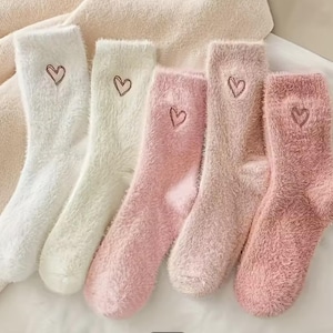 May include: Six pairs of fuzzy socks in shades of white and pink are arranged side by side. Each sock has a small embroidered heart near the top. The socks appear soft and comfortable, perfect for lounging.