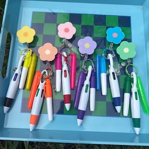 May include: A selection of pens with flower-shaped keyrings in various colours, including yellow, pink, and blue. The pens are white with coloured accents, arranged on a blue tray with a green and black checkered pattern.