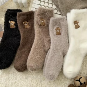 May include: A set of five fuzzy crew socks in black, dark brown, tan, light grey, and white. Each sock features a small, embroidered brown highland cow graphic on the upper ankle area. The socks have a soft, plush, and textured appearance.