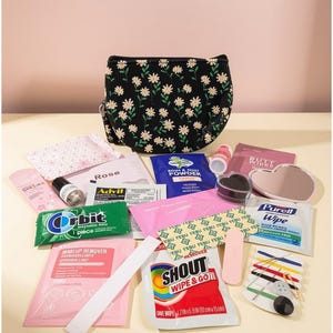 May include: A black crescent-shaped bag with a white daisy pattern sits behind an assortment of items including a pink makeup remover packet, a green Orbit gum pack, a Shout Wipe & Go packet, a heart-shaped mirror, and a small sewing kit.