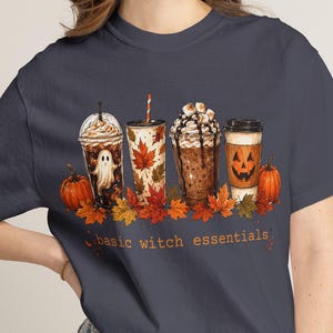 May include: A dark grey short-sleeved t-shirt featuring a graphic of four beverage cups decorated with autumn and Halloween themes, including a ghost, maple leaves, and a jack-o'-lantern, with the text "basic witch essentials" printed below.