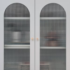 May include: A light grey storage cabinet featuring two doors with arched, vertical reeded glass panels. The cabinet includes two small, round, metallic gold knobs and four slender legs with gold-toned feet, revealing internal shelves.