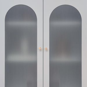 May include: A light grey cabinet with two doors featuring arched, vertical reeded glass panels. The cabinet has two small, round, metallic gold knobs in the centre and stands on four short, thin legs with gold-toned caps at the bottom of each leg.