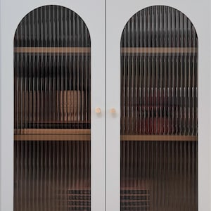 May include: A light grey cabinet with two doors featuring arched, vertical reeded glass panels. The cabinet has small, round, metallic gold knobs and stands on four thin legs with gold-toned feet, revealing internal wooden shelves and objects.