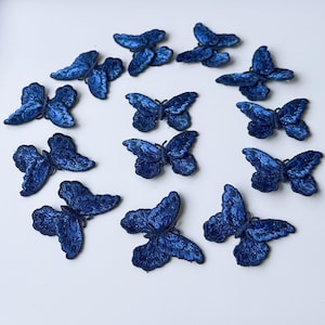 May include: A selection of navy blue embroidered butterfly appliques on a white background. The butterflies, with detailed stitching, are arranged in a circular pattern. These decorative appliques are suitable for various craft projects.