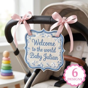 Op de afbeelding: Een crème babybordje met blauwe bloemmotieven en een blauwe rand, met de tekst "Welcome to the world Baby Julian". Het is met roze linten aan een kinderwagen bevestigd. Een stapel kleurrijke houten ringen is zichtbaar op de achtergrond.