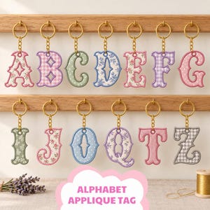 May include: A collection of thirteen embroidered alphabet letter keychains featuring various fabric patterns including floral, gingham, and polka dot designs. Each letter is attached to a gold-toned metal split ring and chain, hanging from a wooden rack.