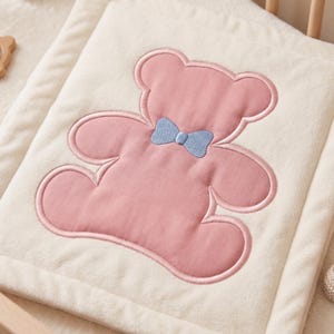 May include: A soft, cream-colored baby blanket with a pink teddy bear design. The bear has a light blue bow tie and is embroidered on the blanket. The blanket is rectangular and has a plush texture.