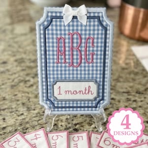 May include: A rectangular fabric milestone marker featuring a blue and white gingham pattern, a white bow at the top, and pink embroidered monogram letters ABG. A white rectangular patch below displays the pink embroidered text 1 month.