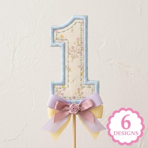 May include: A number one cake topper featuring a cream-colored fabric with a small pink and yellow floral pattern, outlined in light blue stitching. The base is decorated with a layered bow in lavender, dusty rose, and pale yellow with a small rose.