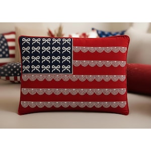 Op de afbeelding: Een rechthoekig rood kussen met een ontwerp dat lijkt op de Amerikaanse vlag. Het blauwe vierkant heeft witte strikaccenten en de rode strepen zijn vervangen door witte kanten details. Een decoratief woonaccessoire.