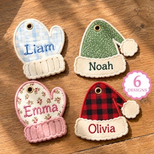 May include: Four embroidered fabric ornaments shaped like mittens and hats with metal eyelets. The items feature names Liam, Noah, Emma, and Olivia in various colours and patterns including blue plaid, green speckled, floral, and red black buffalo check.