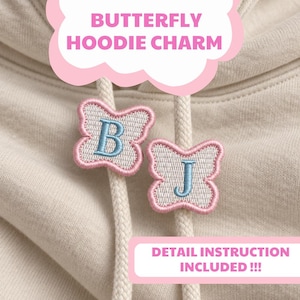 May include: Two butterfly-shaped hoodie charms attached to the drawstrings of a beige hooded sweatshirt. Each charm features a light blue embroidered letter, B and J, surrounded by a white center and a pink stitched border. Text reads BUTTERFLY HOODIE CHARM and DETAIL INSTRUCTION INCLUDED !!!